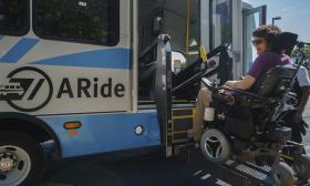 A passenger in a wheelchair is lifted up to ride on the A-Ride bus.